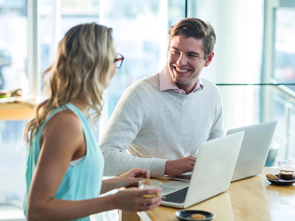 On site training - a male trainer sits next to a female student in a modern office, there is a large window behind them, they both have laptop computers open and are happily in conversation